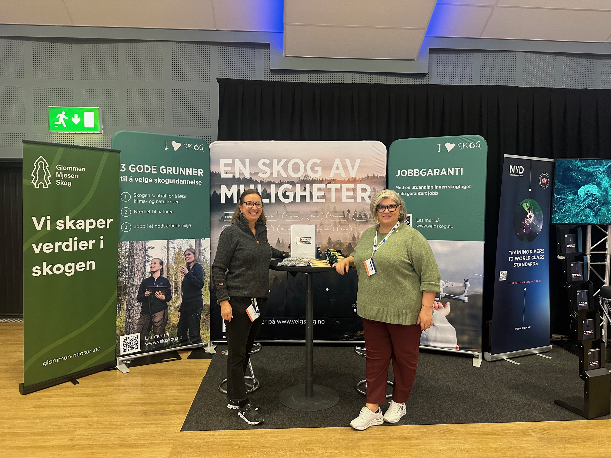 to damer står foran en velg skog stand på en messe, med messevegger bak seg