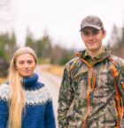 jente med lyst hår og blå strikkegenser og gutt med jaktjakke og jaktcaps står på en grusvei i skogen